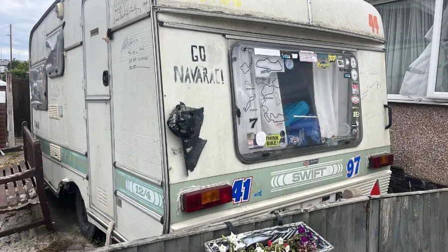 Graffitied touring caravan with a black bin bag stuffed through a broken rear window, wedged against a fence — long past repair and still a priced Bedfordshire collection