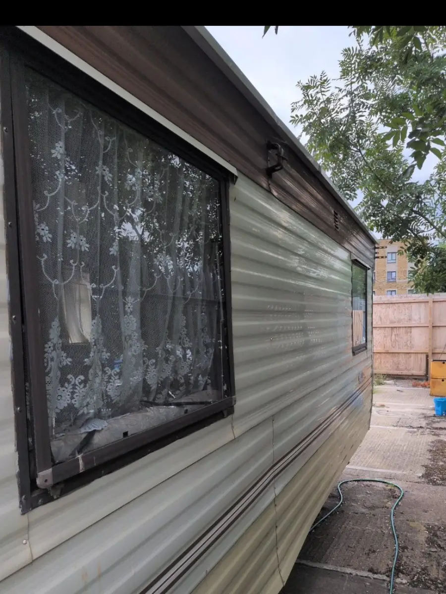 Static caravan flank in corrugated panelling with brown window frames and net curtains still up, standing on concrete hardstanding with a fence tight alongside