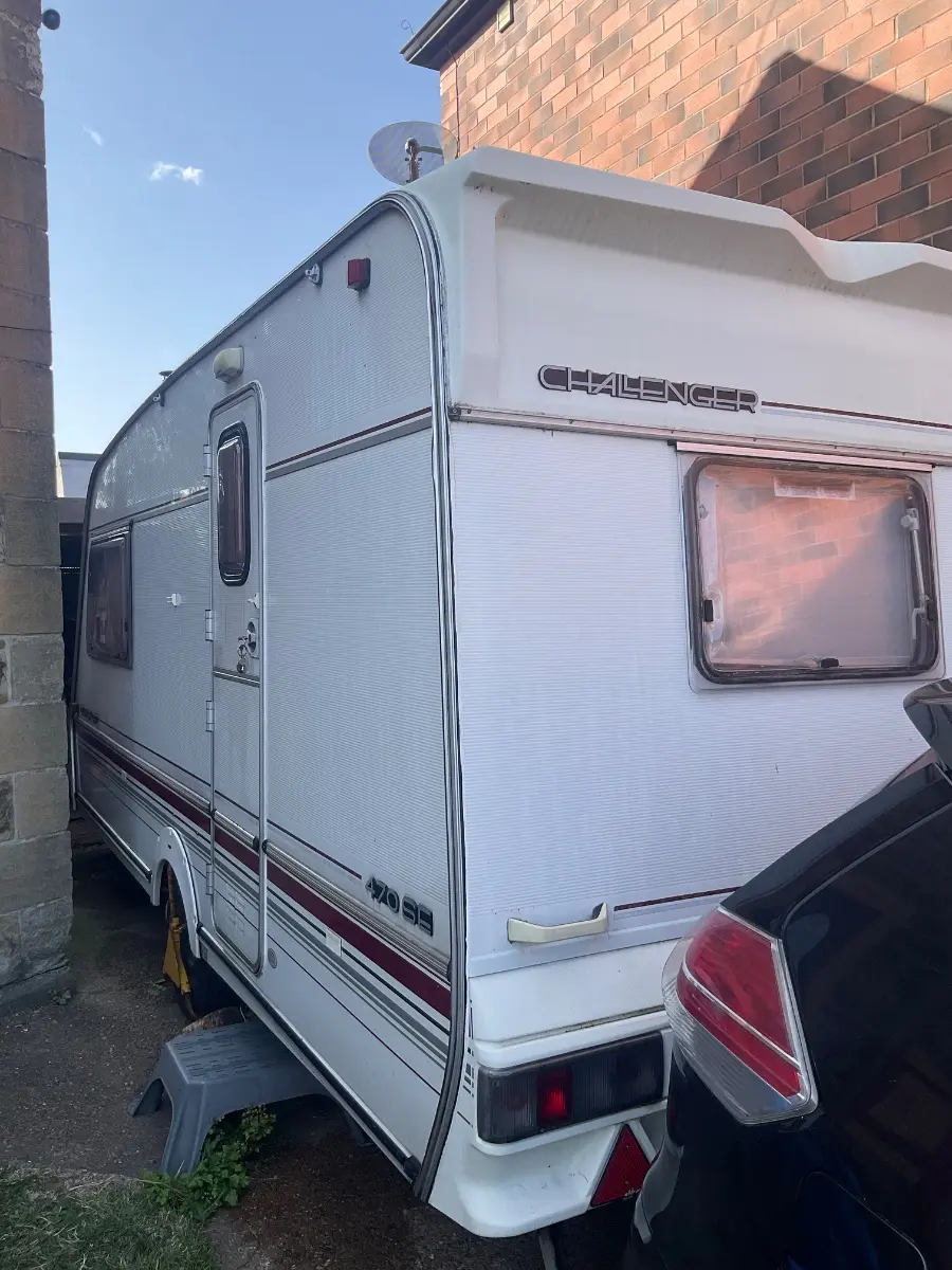 Touring caravan seen from the rear three-quarter on a private driveway beside a brick house, wheels chocked and a car parked behind it