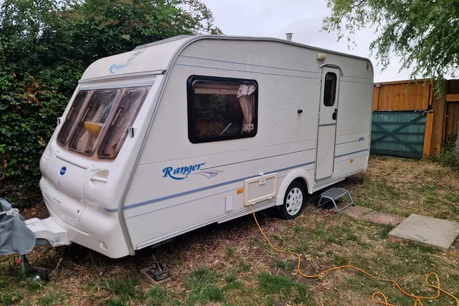 White Bailey Ranger touring caravan standing on rough grass beside a wooden fence, hooked up to an orange extension lead with a chock and a pallet on the ground — the everyday starting point for a Windsor collection