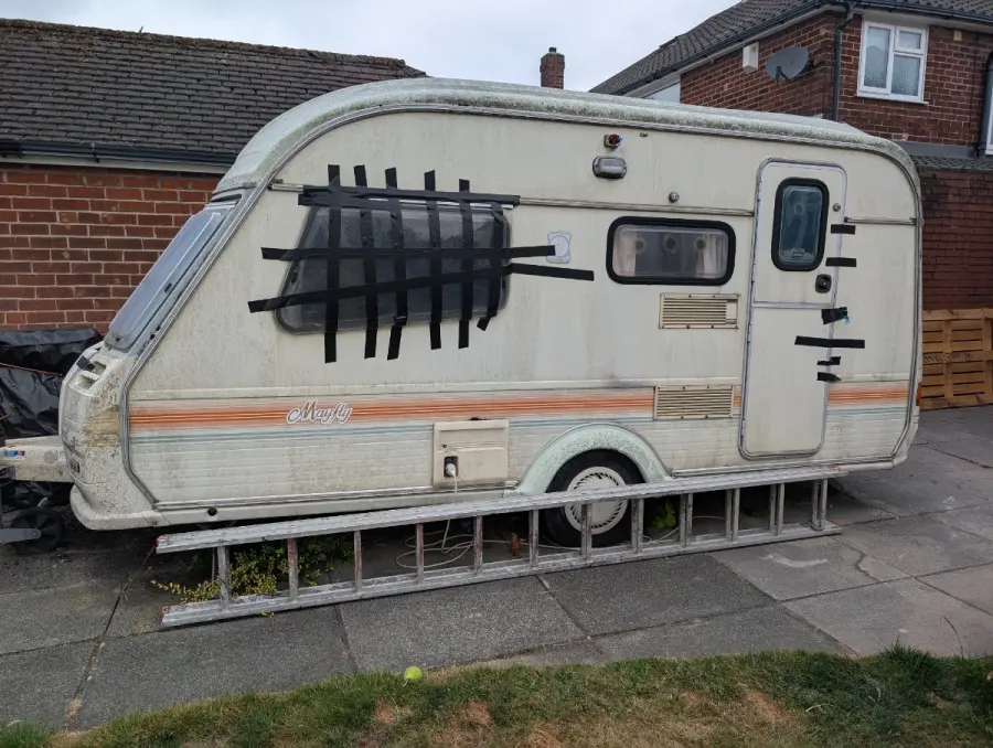 Derelict touring caravan wedged on a suburban driveway beside a brick house, its side window taped over and a ladder propped against the wall — a typical restricted-access collection