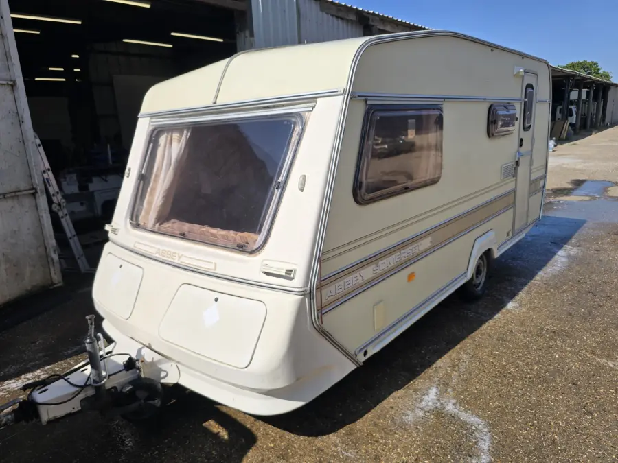 Cream touring caravan standing hitch-forward on the hardstanding outside a workshop, clean and complete — the towable condition that prices at the lower Bedfordshire band