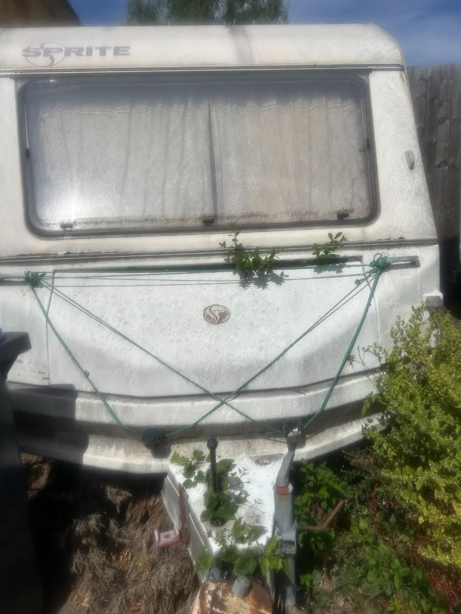 Front of a weathered white touring caravan with its hitch exposed and cover strapped down, ready to be moved once a collection slot is booked