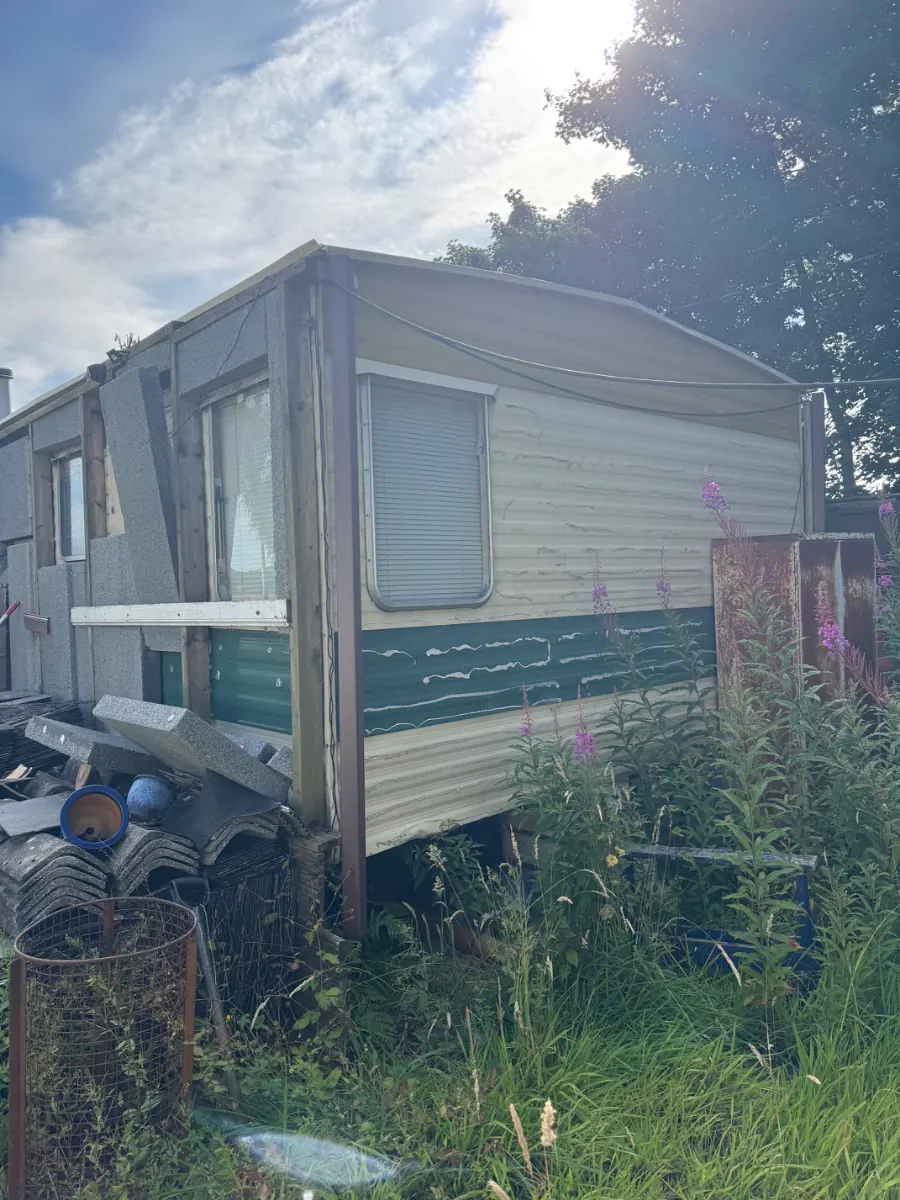 Static caravan standing on blocks in waist-high grass, panels weathered and debris piled underneath, the kind of job a household recycling centre cannot take