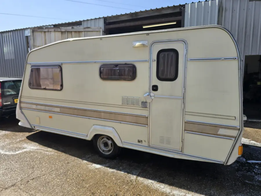 Cream 1980s touring caravan standing outside a corrugated workshop, wheels on and sound enough to be towed away whole