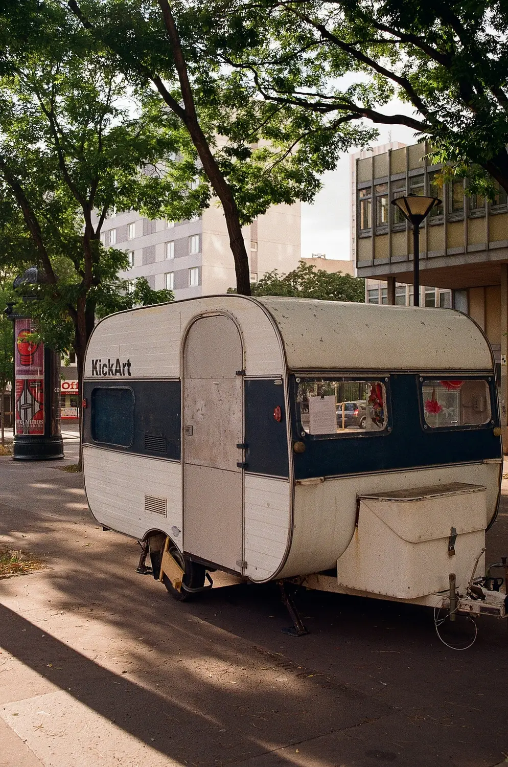 Vintage touring caravan parked kerbside on a tree-lined street between town-centre buildings, the sort of tight urban pitch a Dunstable collection has to reverse out of