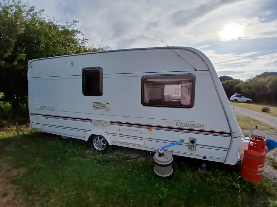 White touring caravan parked on grass beside a hedge with a gas bottle and water barrel alongside, booked in for collection