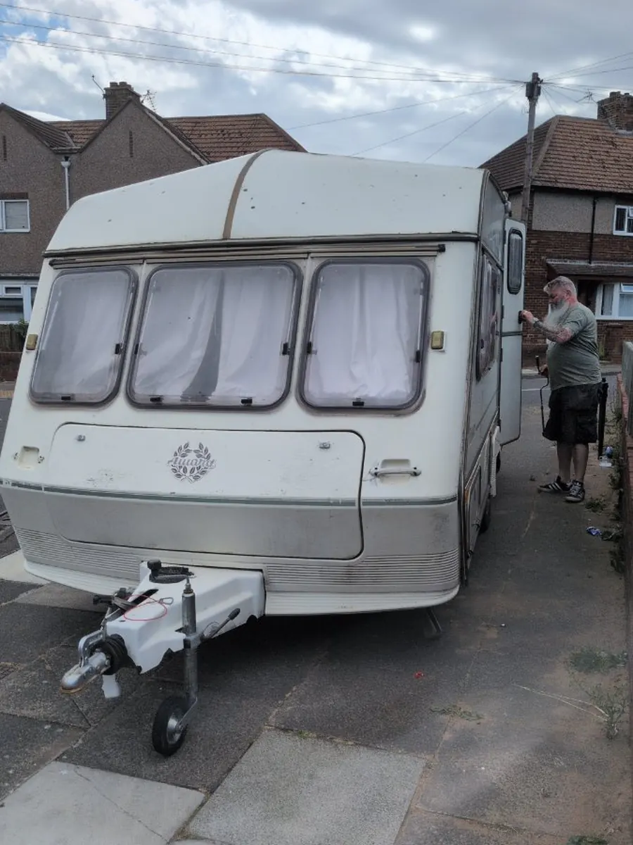 Touring caravan hitched and ready on a suburban driveway with someone standing alongside and the curtains still drawn, the moment before a booked collection