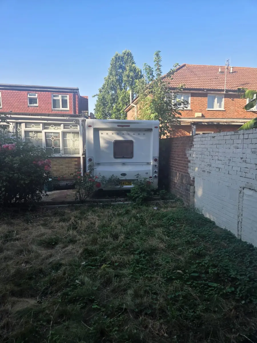Caravan standing at the far end of an overgrown back garden behind a suburban semi, reachable only past a brick side wall, the layout behind a lot of Slough driveway collections
